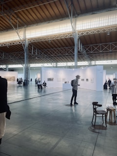 An art exhibition in a large, spacious hall with high ceilings, illuminated by soft lighting from the windows above. White walls partition the space into different sections, each displaying various artworks. Several people are strolling around, observing the art. A few chairs are scattered across the floor.