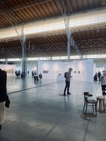 An art exhibition in a large, spacious hall with high ceilings, illuminated by soft lighting from the windows above. White walls partition the space into different sections, each displaying various artworks. Several people are strolling around, observing the art. A few chairs are scattered across the floor.