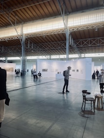 An art exhibition in a large, spacious hall with high ceilings, illuminated by soft lighting from the windows above. White walls partition the space into different sections, each displaying various artworks. Several people are strolling around, observing the art. A few chairs are scattered across the floor.