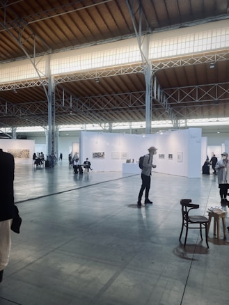 An art exhibition in a large, spacious hall with high ceilings, illuminated by soft lighting from the windows above. White walls partition the space into different sections, each displaying various artworks. Several people are strolling around, observing the art. A few chairs are scattered across the floor.