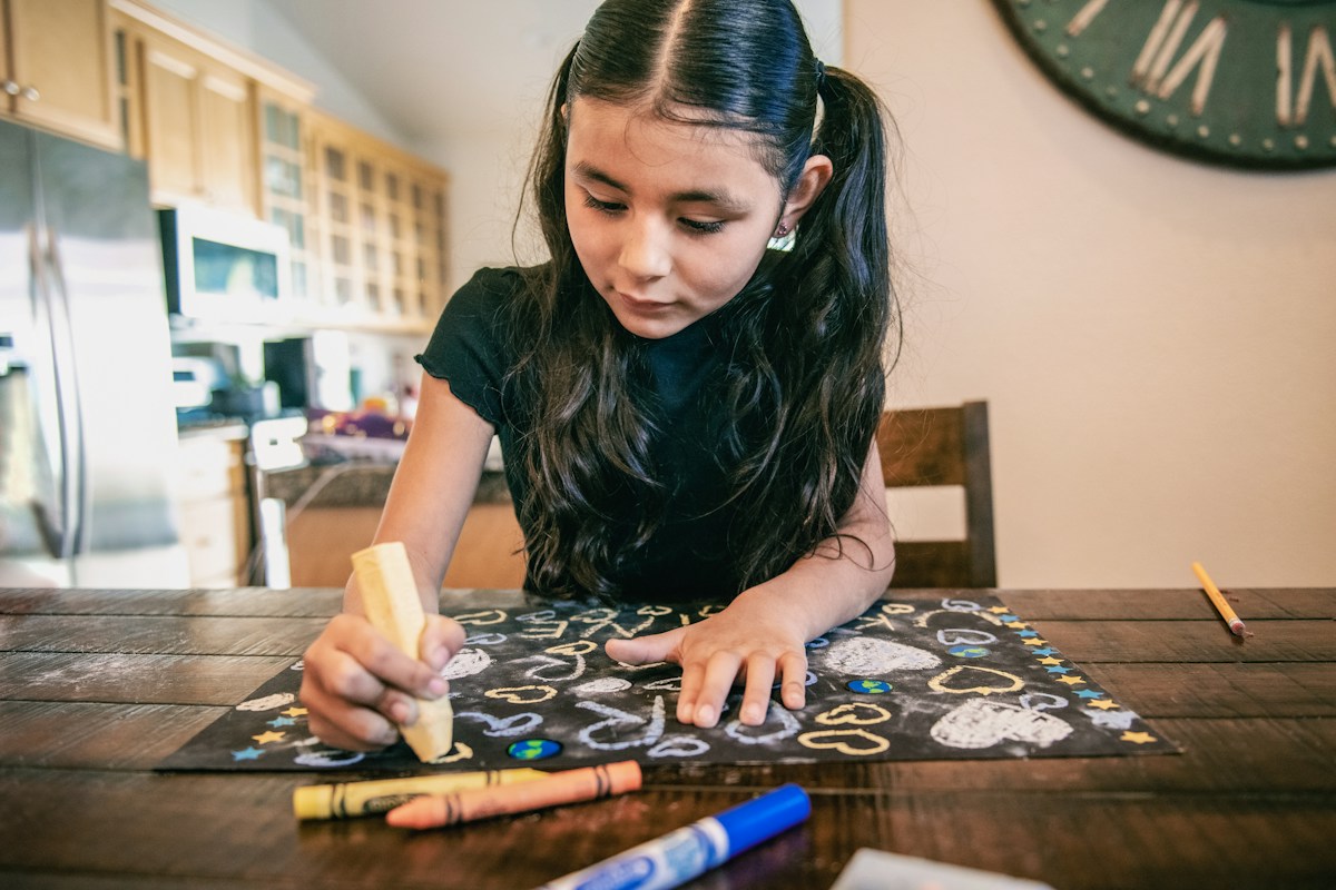 Young girl drawing with crayons at a table, creating a card for Teacher Appreciation Week.