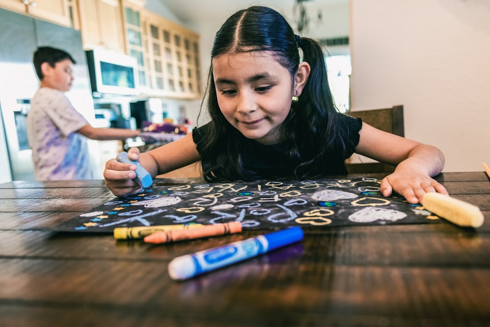 a little girl sitting at a table with crayons and markers