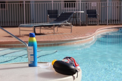 A poolside scene with a bottle of sunscreen on a round table near a swimming pool. The clear blue water of the pool is inviting, with lounge chairs and a metal fence in the background. Sunlight casts shadows, enhancing the summery atmosphere.