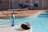 A poolside scene with a bottle of sunscreen on a round table near a swimming pool. The clear blue water of the pool is inviting, with lounge chairs and a metal fence in the background. Sunlight casts shadows, enhancing the summery atmosphere.