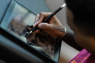 A person with a tattooed hand is using a stylus to draw on a tablet. The artwork on the screen appears detailed and intricate. The focus is on the hand and the digital illustration process.
