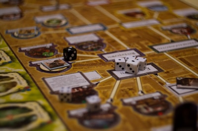 A board game setup with several black and white dice scattered across the board. The board features various sections with printed text and graphics, connected by lines. The focus is on the dice, which are central to the gameplay.