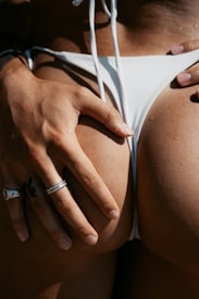 A close-up of a person wearing a white bikini, with a hand resting on their hip. The hand is adorned with rings, and there are water droplets visible on the skin, suggesting a beach or pool setting.