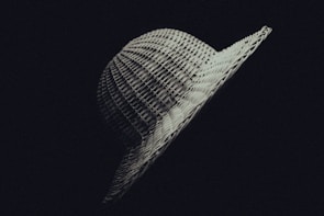 A woven wicker hat with a broad brim is set against a dark background. The intricate pattern of interlacing material creates a textured appearance. The lighting highlights the details and shadows within the weave, giving a three-dimensional quality.