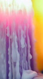 Melted wax drips down a surface with a gradient of colors blending from soft pink to bright yellow. The texture is smooth yet irregular, reminiscent of flowing liquid frozen in time.