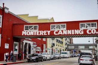 A street scene featuring the historic Monterey Canning Company building with a red and white sign spanning across the street. Cars are parked along the road, and people are walking on the sidewalk near shops and buildings painted in red and yellow.