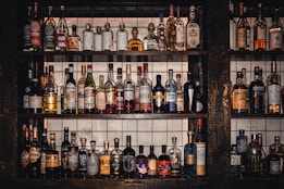 A collection of various liquor bottles of different shapes, sizes, and colors neatly arranged on three rows of dark wooden shelves. The background behind the shelves features white tiles with grid lines. Each bottle has uniquely designed labels, displaying a diverse selection of spirits, including whiskey, tequila, and liqueurs.