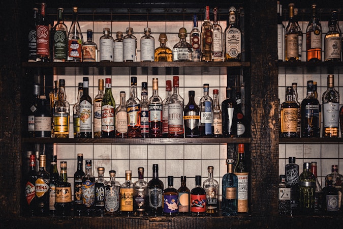 A collection of various liquor bottles of different shapes, sizes, and colors neatly arranged on three rows of dark wooden shelves. The background behind the shelves features white tiles with grid lines. Each bottle has uniquely designed labels, displaying a diverse selection of spirits, including whiskey, tequila, and liqueurs.