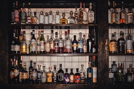 A collection of various liquor bottles of different shapes, sizes, and colors neatly arranged on three rows of dark wooden shelves. The background behind the shelves features white tiles with grid lines. Each bottle has uniquely designed labels, displaying a diverse selection of spirits, including whiskey, tequila, and liqueurs.