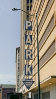 A large vertical sign spelling out the word 'PARK' is mounted on the side of a tall building. The letters are blue with a white background. Below the word 'PARK,' a triangular sign reads 'TOWER GARAGE COMPLETE SERVICE.' The building features a uniform architectural style with a grid-like design. A street lamp and a small 'No Parking Anytime' sign are visible near the bottom.
