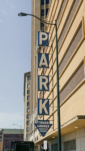 A large vertical sign spelling out the word 'PARK' is mounted on the side of a tall building. The letters are blue with a white background. Below the word 'PARK,' a triangular sign reads 'TOWER GARAGE COMPLETE SERVICE.' The building features a uniform architectural style with a grid-like design. A street lamp and a small 'No Parking Anytime' sign are visible near the bottom.
