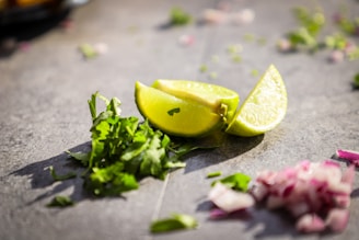 Slices of lime and chopped cilantro are placed on a gray surface, with some diced red onions scattered nearby. The sunlight casts shadows, adding a fresh and vibrant look.