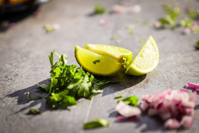 Slices of lime and chopped cilantro are placed on a gray surface, with some diced red onions scattered nearby. The sunlight casts shadows, adding a fresh and vibrant look.