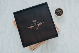 A black box with elegant lettering sits on a soft white surface. The box is labeled 'Wedding Memories' along with the names 'Sagar' and 'Yasha' and a date. To the side, a small potted succulent plant adds a touch of nature.