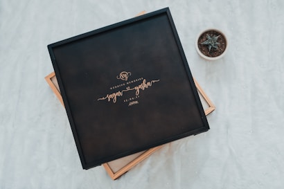 A black box with elegant lettering sits on a soft white surface. The box is labeled 'Wedding Memories' along with the names 'Sagar' and 'Yasha' and a date. To the side, a small potted succulent plant adds a touch of nature.