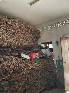 A large stack of firewood is neatly piled up against a wall inside a room. Above the pile, there are some bags and miscellaneous items, and a small window can be seen in the background. A partially open wooden door is visible, along with a green shopping bag hanging on a handle.