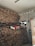 A large stack of firewood is neatly piled up against a wall inside a room. Above the pile, there are some bags and miscellaneous items, and a small window can be seen in the background. A partially open wooden door is visible, along with a green shopping bag hanging on a handle.