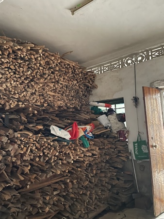 A large stack of firewood is neatly piled up against a wall inside a room. Above the pile, there are some bags and miscellaneous items, and a small window can be seen in the background. A partially open wooden door is visible, along with a green shopping bag hanging on a handle.