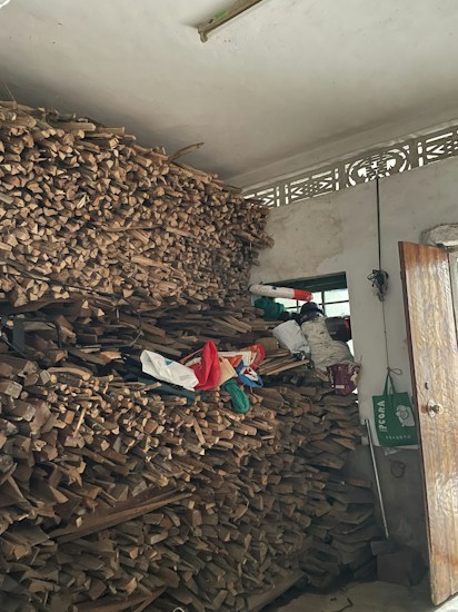 A large stack of firewood is neatly piled up against a wall inside a room. Above the pile, there are some bags and miscellaneous items, and a small window can be seen in the background. A partially open wooden door is visible, along with a green shopping bag hanging on a handle.