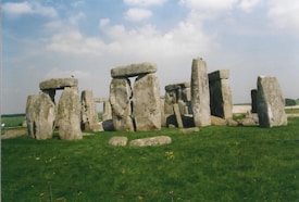 A historical arrangement of large standing stones, known as Stonehenge, is set on a verdant green field under a partly cloudy sky. The stones form a circular pattern, with some horizontal lintels connecting pairs of upright stones.