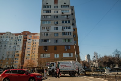 A multi-story residential building stands against a clear blue sky. A garbage truck with text indicating a city cleanliness initiative is parked in front of the building. A red car is parked nearby, and a person is seen walking on the sidewalk.