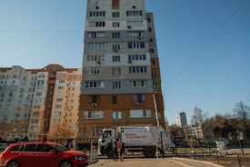 A multi-story residential building stands against a clear blue sky. A garbage truck with text indicating a city cleanliness initiative is parked in front of the building. A red car is parked nearby, and a person is seen walking on the sidewalk.