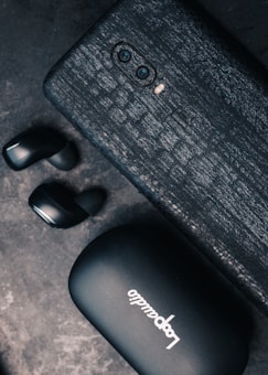 A textured black smartphone with dual cameras is placed next to a pair of black wireless earbuds. The earbuds rest near a black case with the brand name 'LoopAudio' visible in white lettering.