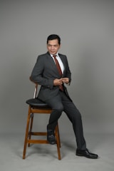 A man in a gray suit and white shirt is seated on a wooden stool. He is wearing a maroon tie and black shoes, and his posture is slightly turned to the side with a confident expression.