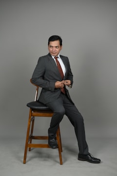 A man in a gray suit and white shirt is seated on a wooden stool. He is wearing a maroon tie and black shoes, and his posture is slightly turned to the side with a confident expression.