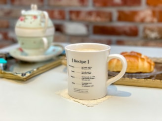 A ceramic mug with a recipe printed on it sits on a coaster. In the background, there's a floral-patterned tea set on a tray and a croissant on a plate. The setting is cozy with a brick wall partially visible behind the objects.