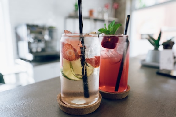 Two refreshing beverages served in clear glasses, each topped with dark straws. The drink on the left is a clear liquid infused with slices of strawberries and cucumbers, while the one on the right is a reddish beverage garnished with a mint sprig and a cherry.