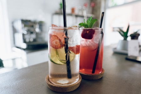 Two refreshing beverages served in clear glasses, each topped with dark straws. The drink on the left is a clear liquid infused with slices of strawberries and cucumbers, while the one on the right is a reddish beverage garnished with a mint sprig and a cherry.