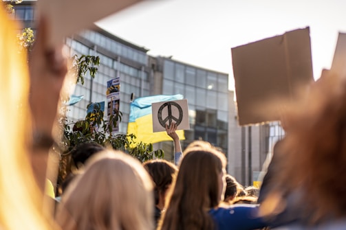 A group of people participate in a protest or rally. One hand holds a sign with a peace symbol. There is a Ukrainian flag visible, and the setting is against a backdrop of modern buildings. The atmosphere is lively and filled with people holding various signs.