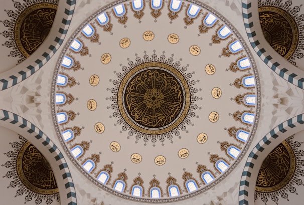 An intricately designed dome featuring elaborate calligraphy and geometric patterns. The design is symmetrical with repeated motifs and colored elements, including blue, gold, and white hues. The calligraphy appears to be Arabic and is central to the aesthetic of the dome.