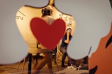 A person is sitting in a thoughtful pose inside a cozy, warmly lit room. The scene is viewed through a large heart-shaped cutout on glass. Another person is walking by, partially obscured by the heart shape. The interior is decorated with small images on the wall.