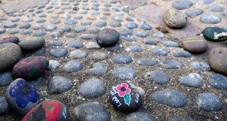 Painted stones are scattered atop a pebble-covered surface. Some of the stones have flowers and other designs painted on them in various colors.