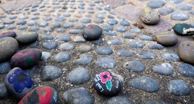 Painted stones are scattered atop a pebble-covered surface. Some of the stones have flowers and other designs painted on them in various colors.