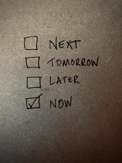 A handwritten checklist on a textured surface with four items labeled 'Next', 'Tomorrow', 'Later', and 'Now', with 'Now' being checked.