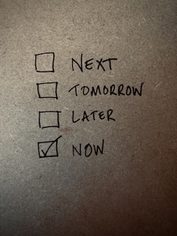 A handwritten checklist on a textured surface with four items labeled 'Next', 'Tomorrow', 'Later', and 'Now', with 'Now' being checked.
