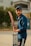 A young man stands outside holding a cricket bat, wearing a blue shirt and cap. Behind him, there's a house and green foliage.