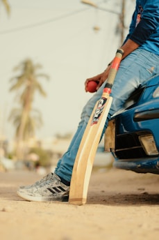 A person in jeans and a blue shirt is sitting on the front bumper of a parked blue car, holding a red cricket ball. A cricket bat is resting against the ground. A blurred background shows palm trees and a sandy ground.