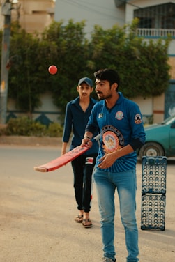 Two individuals are engaged in a casual cricket session on a street. One person in a blue sports jersey is poised to hit a cricket ball with a bat. Another individual stands behind, wearing casual attire with a cap. A stack of blue crates serves as the makeshift wickets. Trees and a building are visible in the background, adding to the outdoor setting.