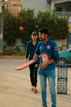 Two individuals are engaged in a casual cricket session on a street. One person in a blue sports jersey is poised to hit a cricket ball with a bat. Another individual stands behind, wearing casual attire with a cap. A stack of blue crates serves as the makeshift wickets. Trees and a building are visible in the background, adding to the outdoor setting.