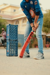 A person wearing a blue sports jersey and jeans is holding an orange cricket bat, appearing ready to play. To the left, there are two stacks of blue Pepsi crates arranged vertically. The background shows a residential building and some greenery.
