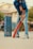 A person wearing a blue sports jersey and jeans is holding an orange cricket bat, appearing ready to play. To the left, there are two stacks of blue Pepsi crates arranged vertically. The background shows a residential building and some greenery.