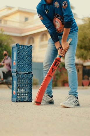A person wearing a blue sports jersey and jeans is holding an orange cricket bat, appearing ready to play. To the left, there are two stacks of blue Pepsi crates arranged vertically. The background shows a residential building and some greenery.